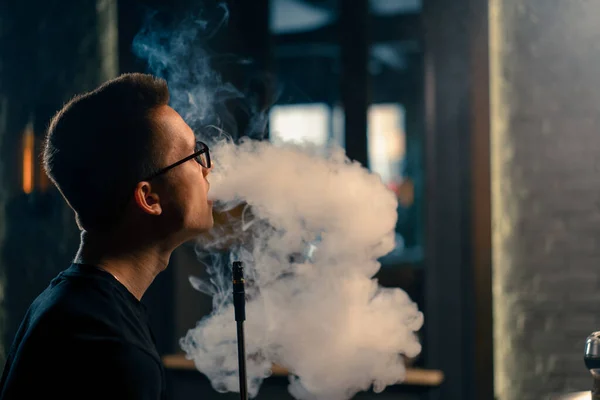 A hookah man in glasses smokes a traditional hookah pipe A man exhales smoke in hookah cafe or lounge bar close-up