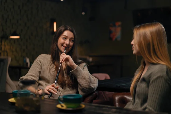 two young female friends are sitting relaxed in hookah bar smoking hookah and having fun chatting recreation concept