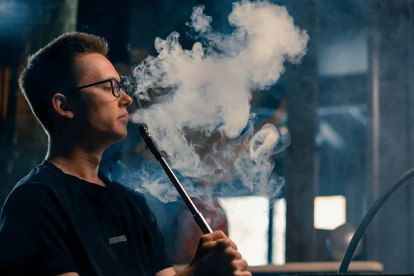 A man in glasses smokes a traditional hookah pipe A man exhales thick smoke in hookah cafe or lounge bar