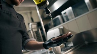 Otel restoranında profesyonel mutfak, şef bir hamburger pirzolasına yağ döker ve elleriyle yağlar.