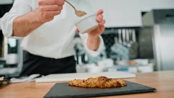 プロのキッチンで料理をするシェフが焼いたチキンフィレの上に蜂蜜マスタードソースをかけるパンくずリスト — ストック動画