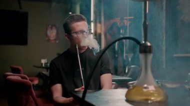 Gözlüklü bir adam geleneksel nargile piposu tüttürür. Bir adam nargile kafeteryasında ya da bar barında yoğun duman çıkarır.