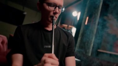 Gözlüklü bir adam geleneksel nargile piposu tüttürür. Bir adam nargile kafeteryasında ya da bar barında yoğun duman çıkarır.