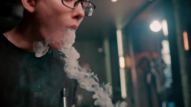 Gözlüklü bir adam geleneksel nargile piposu tüttürür. Bir adam nargile kafesinde ya da dinlenme odasında kalın dumanlı yüzükler solur.