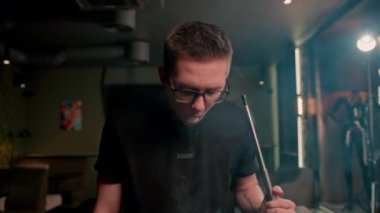 Gözlüklü bir adam geleneksel nargile piposu tüttürür. Bir adam nargile kafesinde ya da dinlenme odasında kalın dumanlı yüzükler solur.