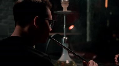 Gözlüklü bir adam geleneksel nargile piposu tüttürür. Bir adam nargile kafeteryasında ya da bar barında yoğun duman çıkarır.