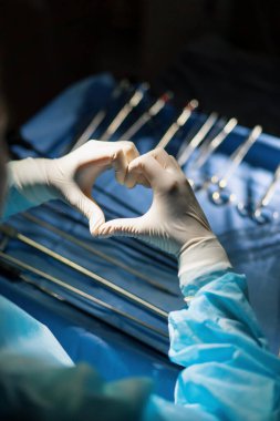 Doktor ameliyat sonrası kalp işaretini elleriyle gösteriyor. Cerrahi gereçlerin arka planında.