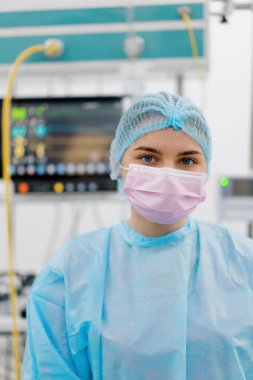 Ameliyat sonrası ameliyathanede maskeli ve steril giysili genç bir bayan doktorun portresi duruyor.