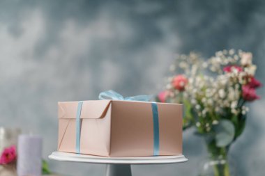 Çiçeklerin arkasında duran fiyonklu bir hediye kutusunda paketlenmiş lezzetli bir pasta.
