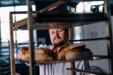 Rafların arasındaki erkek fırıncı fırın endüstrisinde yeni pişmiş ekmeği dikkatle kontrol ediyor.