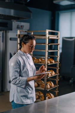 Rafların yanında üniformalı bir kadın fırıncı elinde tabletle ekmek fırınını kontrol ediyor.
