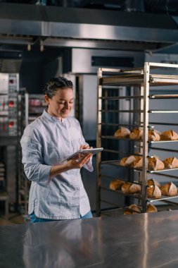 Rafların yanında üniformalı bir kadın fırıncı elinde tabletle ekmek fırınını kontrol ediyor.