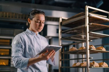 Rafların yanında üniformalı bir kadın fırıncı elinde tabletle ekmek fırınını kontrol ediyor.