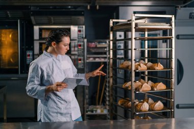 Rafların yanında üniformalı bir kadın fırıncı elinde tabletle ekmek fırınını kontrol ediyor.
