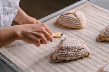 Kadın fırıncı, fırın yapımı hamur işlerini kapatmadan önce, profesyonel bir fırın bıçağıyla çiğ ekmek rulolarında desenler kesiyor.