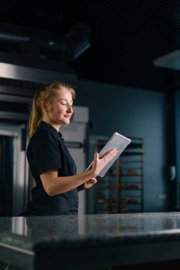 Üniformalı profesyonel bir şekerci elinde tabletle profesyonel mutfakta çalışmaya başlamadan önce görevleri kontrol eder.