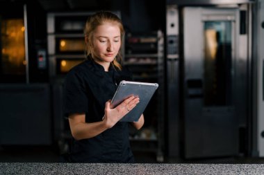 Üniformalı profesyonel bir şekerci elinde tabletle profesyonel mutfakta çalışmaya başlamadan önce görevleri kontrol eder.