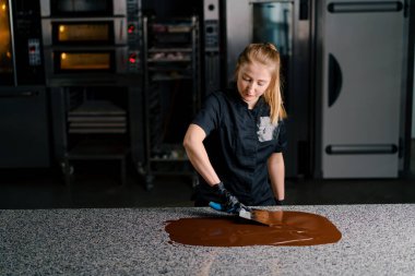 Çikolata üretimini yumuşatan pasta şefi, erimiş çikolatayı hamur işi masasında metal bir spatulayla karıştırıyor.