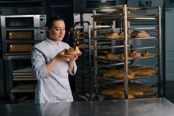 魅力的な女性ベイカーは 棚から新鮮な香りのパンを取ります それは品質ベーカリーのプロのキッチンベーキングをチェックします — ストック写真