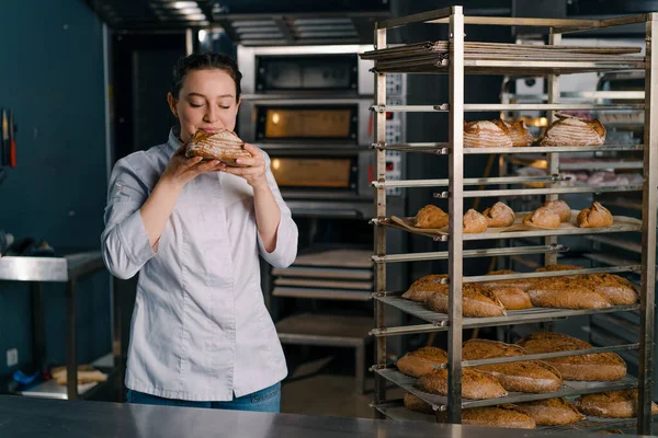 魅力的な女性ベイカーは 棚から新鮮な香りのパンを取ります それは品質ベーカリーのプロのキッチンベーキングをチェックします — ストック写真