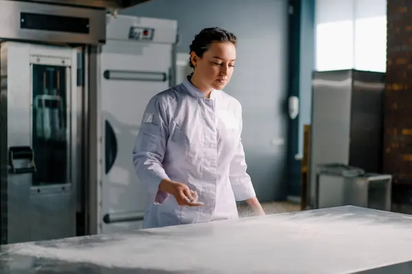 専門のシェフはパン屋の専門の台所でパンのペストリーを準備する前に小麦粉の雲を作るテーブルの上に粉を投げます — ストック写真