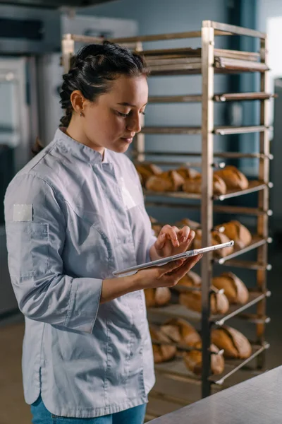 新鮮な焼きパンの棚の近くのユニフォームの女性パン屋は 彼女の手でタブレットをチェックパンパンベーカリー — ストック写真