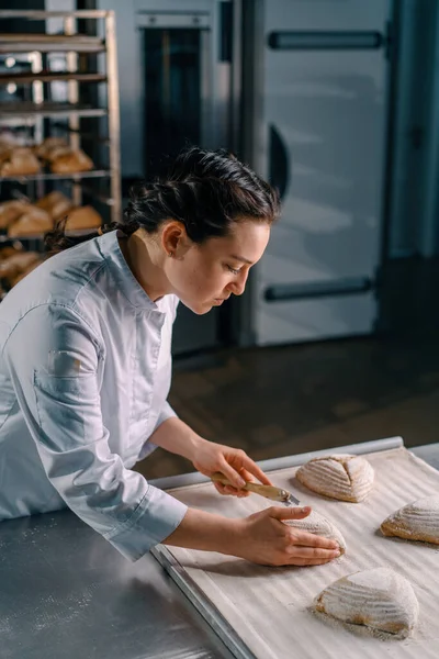 美しい女性のパン屋はパンパンパンにプロのパンナイフでパターンをカットし パン生産のペストリーを焼く前に — ストック写真