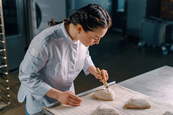 美しい女性のパン屋はパンパンパンにプロのパンナイフでパターンをカットし パン生産のペストリーを焼く前に — ストック写真