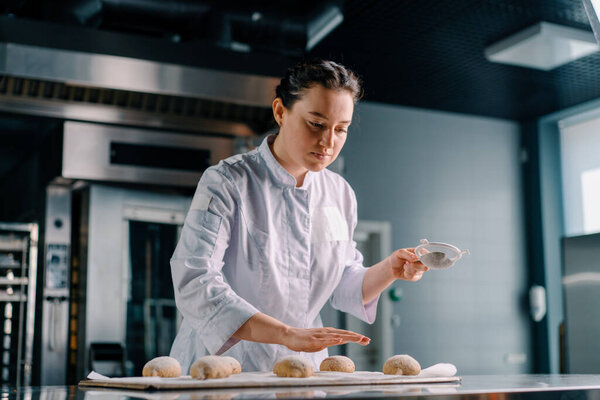 concentrated beautiful woman baker sprinkles flour through sieve raw bread fresh dough before baking baking production bakery
