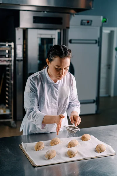 ベーキング生産ベーカリーを焼く前に生パン新鮮な生地をふるうことにより濃縮された美しい女性ベーカースパイク小麦粉 — ストック写真