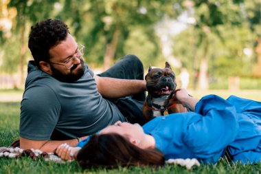 Evli bir çift doğada dinleniyor. Parkta bir çarşafta yatıyor. Fransız buldog cinsinden küçük bir köpekle oynuyor.