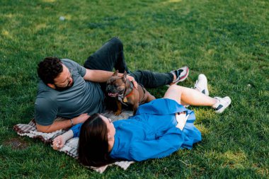 Evli bir çift doğada dinleniyor. Parkta bir çarşafta yatıyor. Fransız buldog cinsinden küçük bir köpekle oynuyor.
