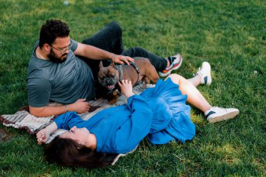 Evli bir çift doğada dinleniyor. Parkta bir çarşafta yatıyor. Fransız buldog cinsinden küçük bir köpekle oynuyor.