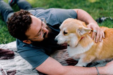 Sakallı adam evcil Corgi köpeğiyle parkta dinleniyor. Yaz pikniğinde yatıyor.