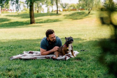 Sakallı bir adam köpeğiyle parkta dinleniyor. Fransız bulldog 'u dışarıda, çarşaf üzerinde piknikte yatıyor.