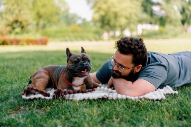 Sakallı bir adam köpeğiyle parkta dinleniyor. Fransız bulldog 'u dışarıda, çarşaf üzerinde piknikte yatıyor.