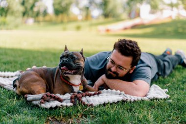Sakallı bir adam köpeğiyle parkta dinleniyor. Fransız bulldog 'u dışarıda, çarşaf üzerinde piknikte yatıyor.
