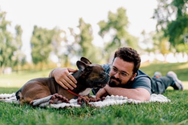 Sakallı bir adam köpeğiyle parkta dinleniyor. Fransız bulldog 'u dışarıda, çarşaf üzerinde piknikte yatıyor.