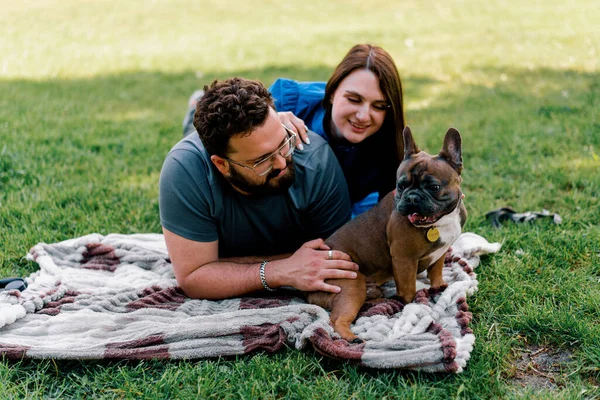 a married couple rests in nature lies on a sheet in the park plays with a small dog of French bulldog breed