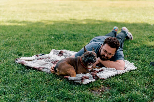 Sakallı bir adam köpeğiyle parkta dinleniyor. Fransız bulldog 'u dışarıda, çarşaf üzerinde piknikte yatıyor.