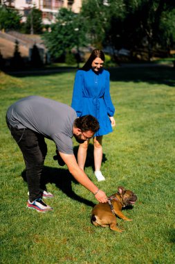 Bir aile, parkta bir buldog köpeğiyle yürüyüşe çıkmış. Sahipleri küçük sevimli bir köpekle oynuyorlar.
