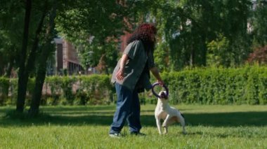 Genç bir kız parkta büyük bir pitbull cinsinin beyaz köpeğiyle oynuyor. Köpek onun peşinden koşuyor ve oyuncağını dişleriyle çekiyor.