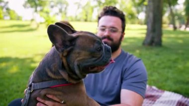 Evli bir çift doğada dinleniyor. Parkta bir çarşafta yatıyor. Fransız buldog cinsinden küçük bir köpekle oynuyor.