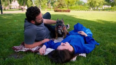 Evli bir çift doğada dinleniyor. Parkta bir çarşafta yatıyor. Fransız buldog cinsinden küçük bir köpekle oynuyor.