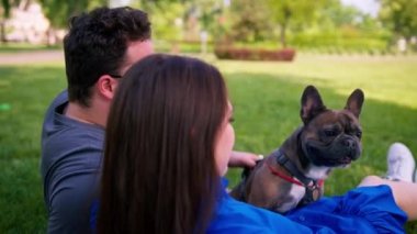 Evli bir çift doğada dinleniyor. Parkta bir çarşafta yatıyor. Fransız buldog cinsinden küçük bir köpekle oynuyor.