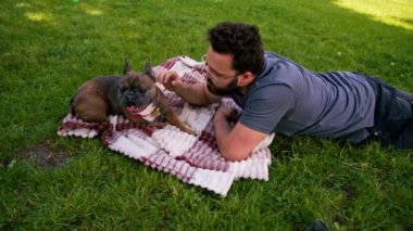 Sakallı bir adam köpeğiyle parkta dinleniyor. Fransız bulldog 'u dışarıda, çarşaf üzerinde piknikte yatıyor.