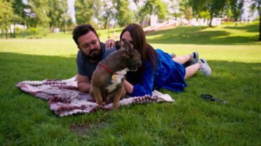 Evli bir çift doğada dinleniyor. Parkta bir çarşafta yatıyor. Fransız buldog cinsinden küçük bir köpekle oynuyor.