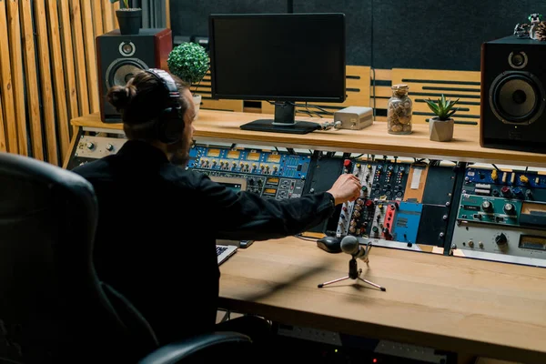年轻的音响工程人员 带着耳机 在有监视器的音乐工作室里工作 还有一个混合主控曲目的均衡器 — 图库照片