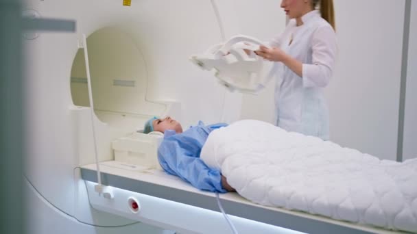 診療所の放射線技師は脳磁気共鳴画像法の前に患者の頭に装置を置きます — ストック動画