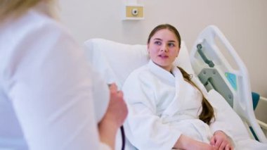 Bir kadın doktor hastane odasında yatağın yanında duruyor ve genç bir kadın hastayla tedavi planını tartışıyor.